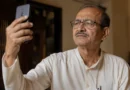 Elderly Indian father holding smartphone slightly above eye level, trying to see screen clearly Title: